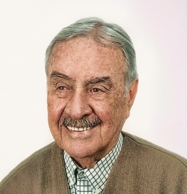 Foto de Alexandre Beldi Netto, senhor com pele branca com sardas, cabelos grisalhos, sobrancelhas arqueadas, olhos pequenos amendoados, nariz grande, lábios grossos em sorriso largo e bigode grisalho. Usa suéter bege sobre camisa quadriculada clara.