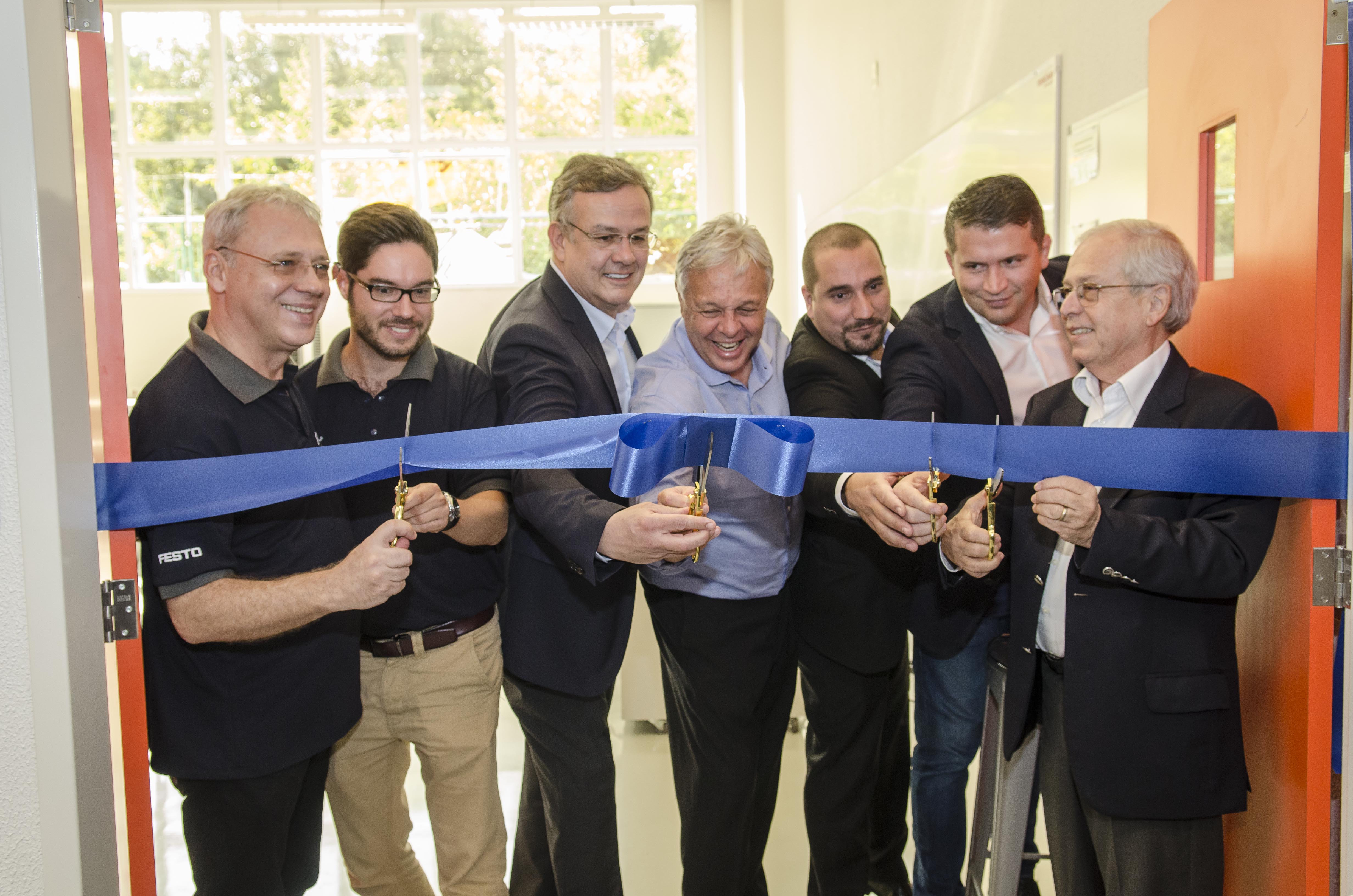 2018. inauguração do laboratório de manufatura avançada. Foto. Sete pessoas sorridentes cortam a faixa inaugural azul.