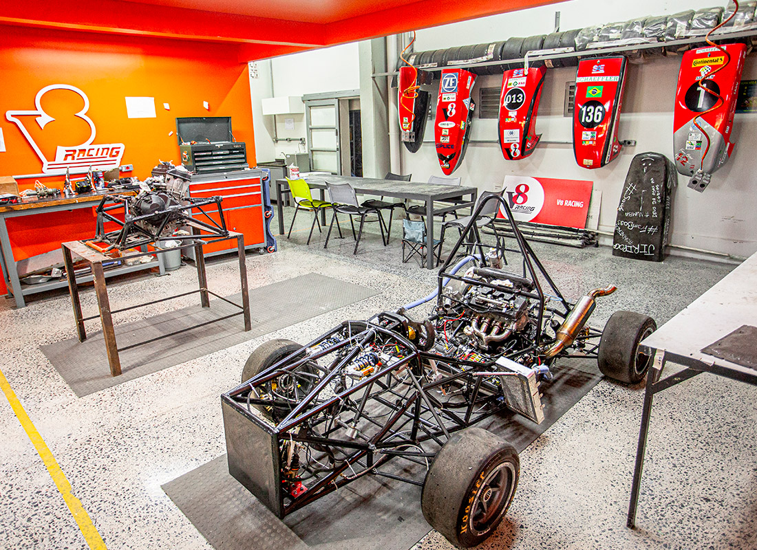 Laboratório de inovação e competições estudantis. Em tons claros e vermelho alaranjado. Bancadas e mesa de trabalho, sobre o piso claro, chassis de carro de corrida e nas paredes carenagens vermelhas penduradas e numeradas.