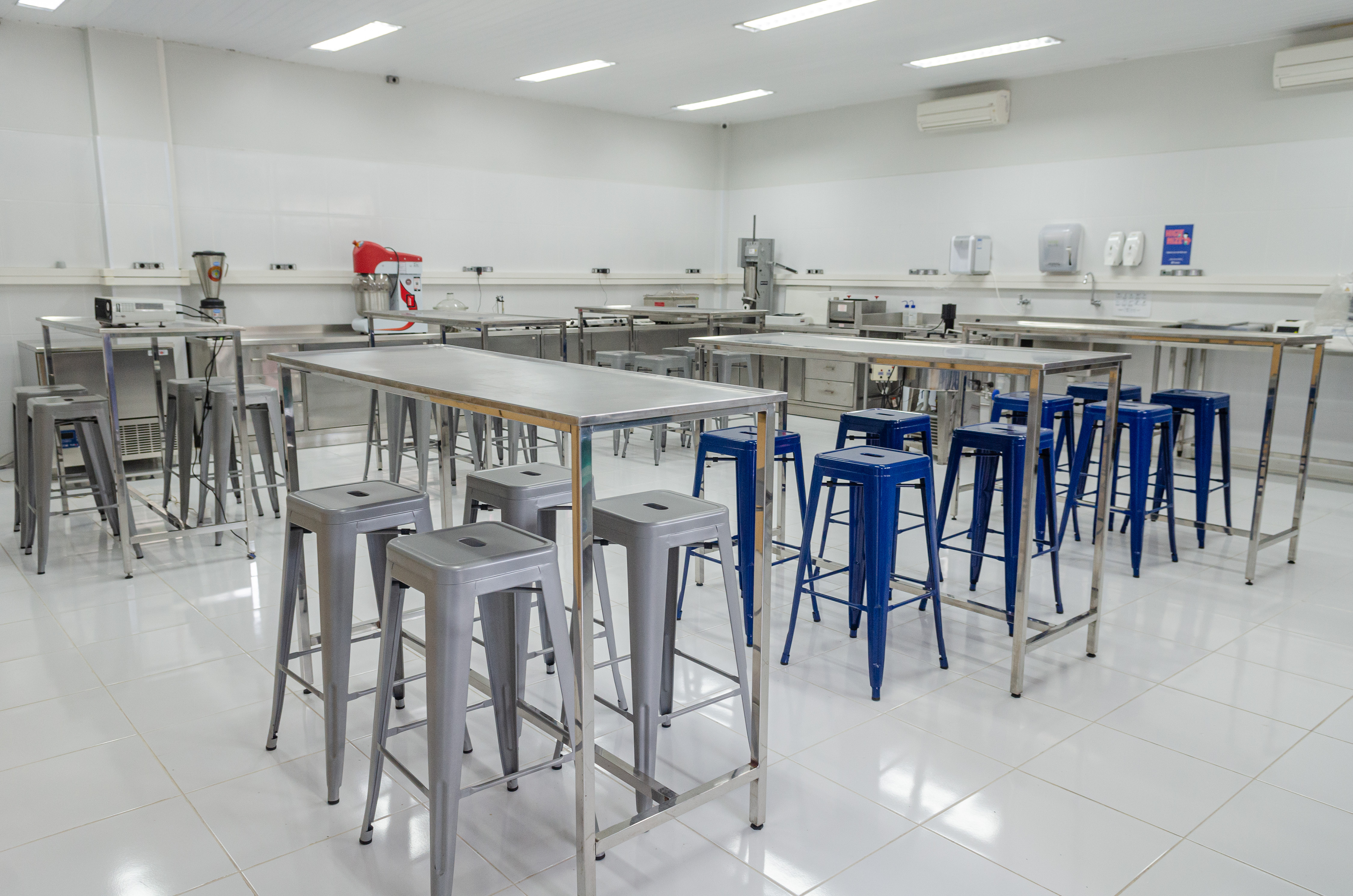 Laboratório de alimentos em tons de branco com mesas altas e banquetas altas ao redor, algumas azuis.