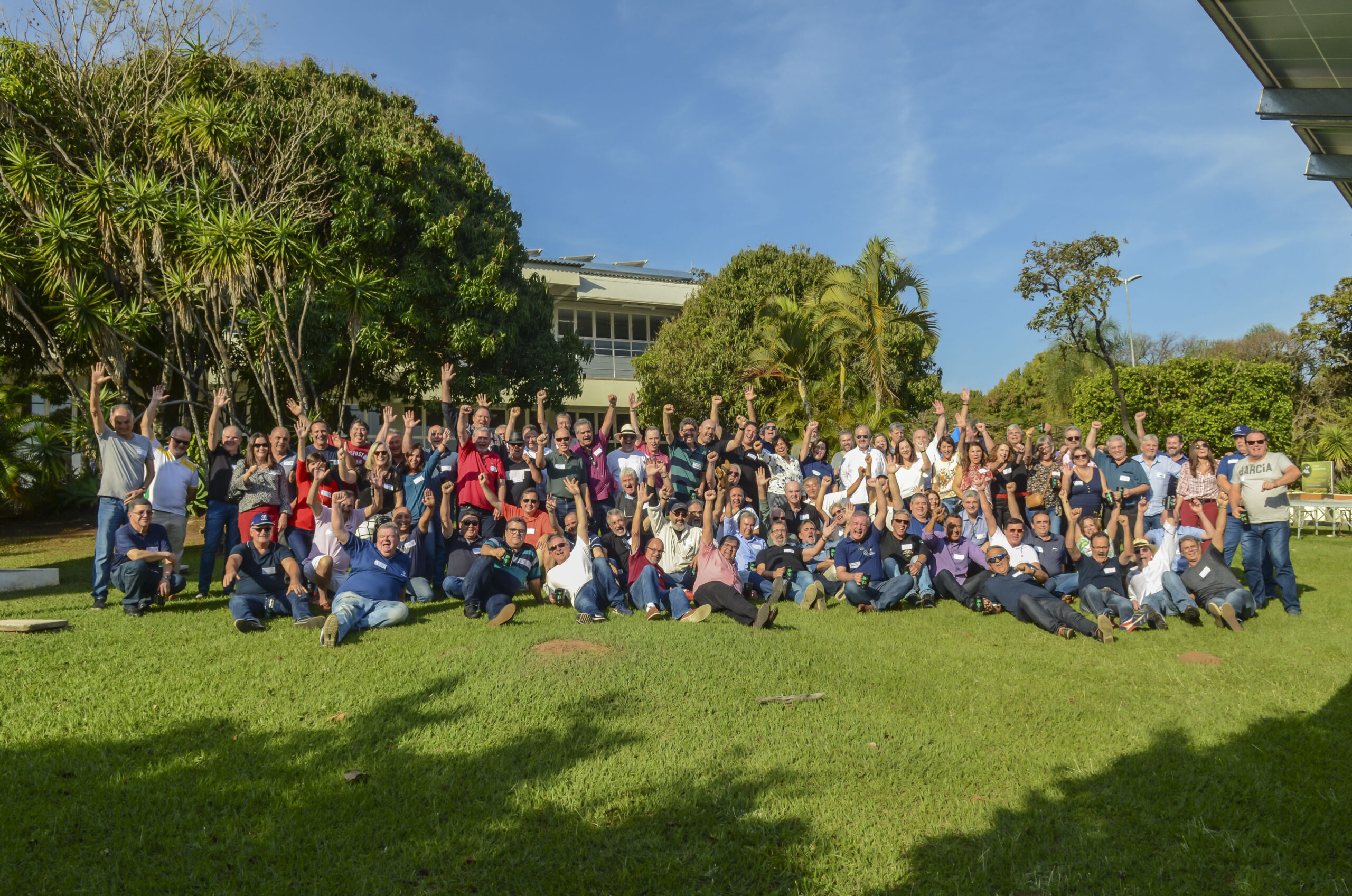 Foto no Campus da Facens. Em dia ensolarado, um grande grupo de alunos e professores reunidos no gramado, alguns sentados outros em pé, posam para a foto com os braços erguidos.