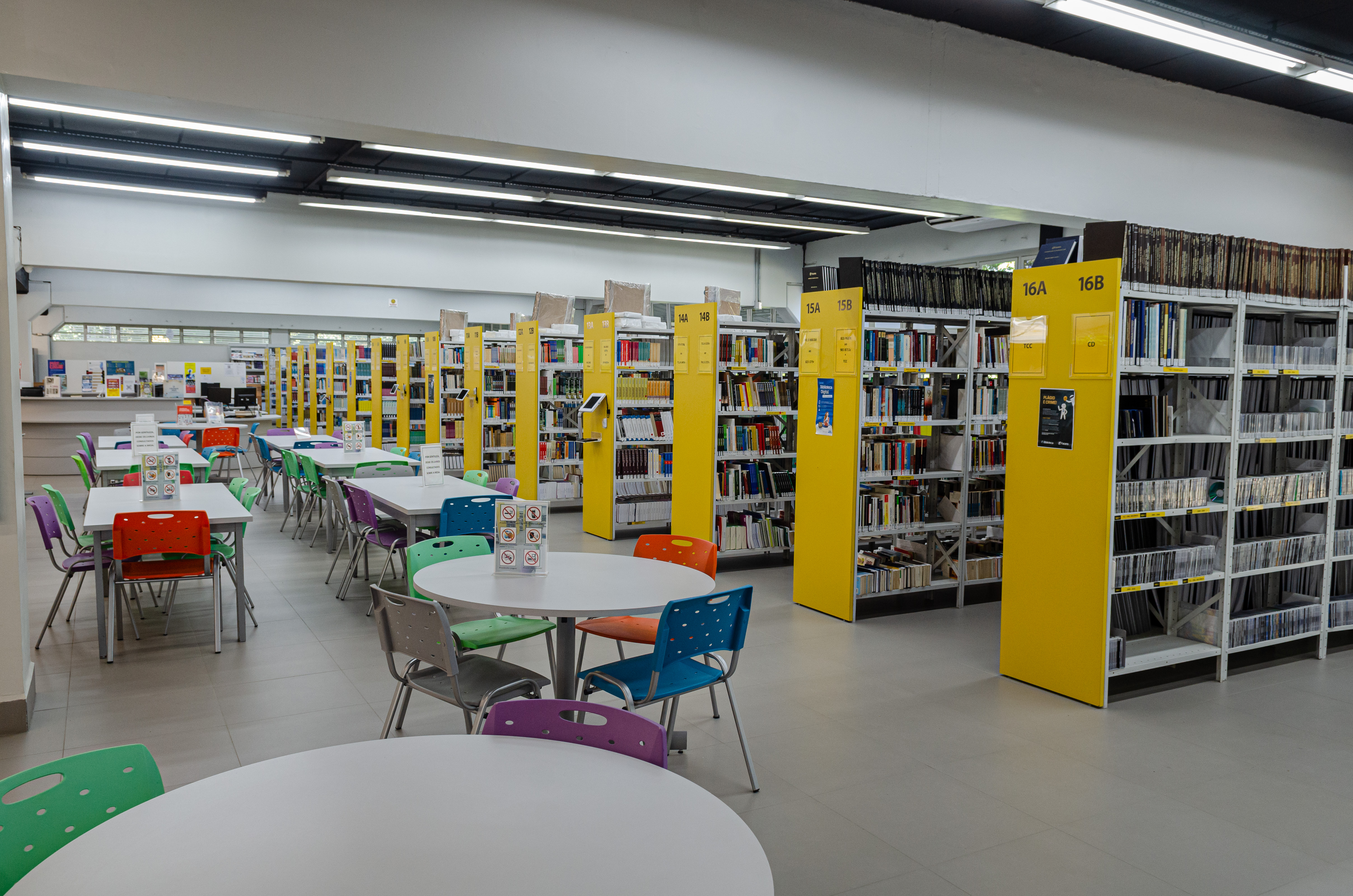 Foto da Biblioteca em ambiente claro composto por mesas redondas rodeadas por cadeiras coloridas. As estantes enfileiradas são amarelas.